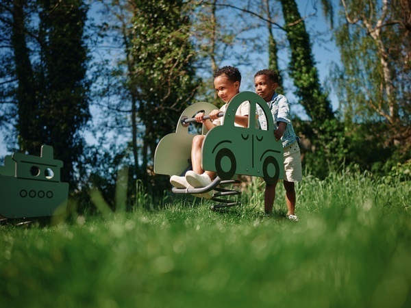 Twee kinderen spelen op een veerelement in de vorm van een auto op een grasveld met bomen op de achtergrond.